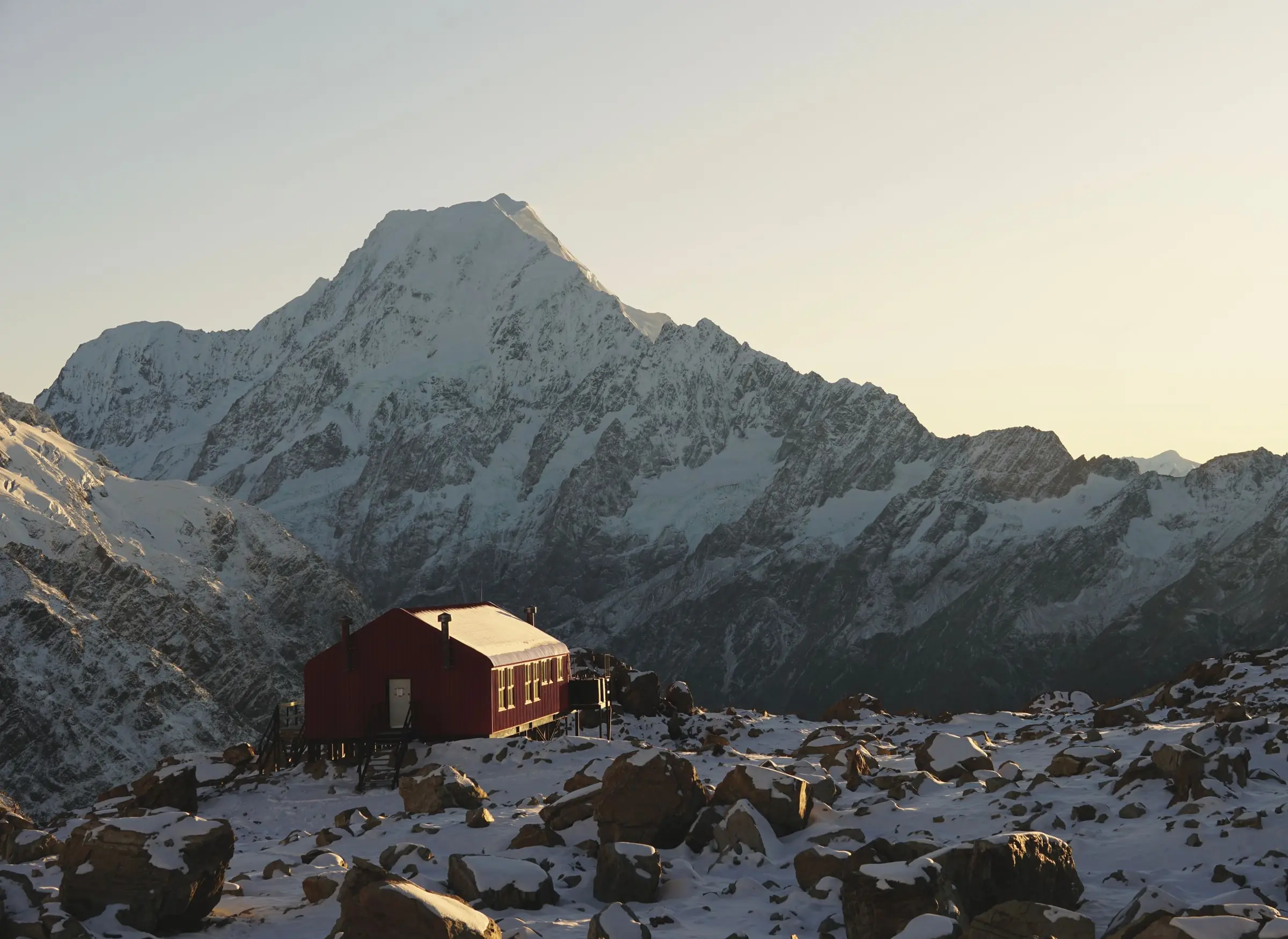 A red alpine hut on a snowy, rocky ridge, with a huge snow-covered mountain face behind it in late light.
