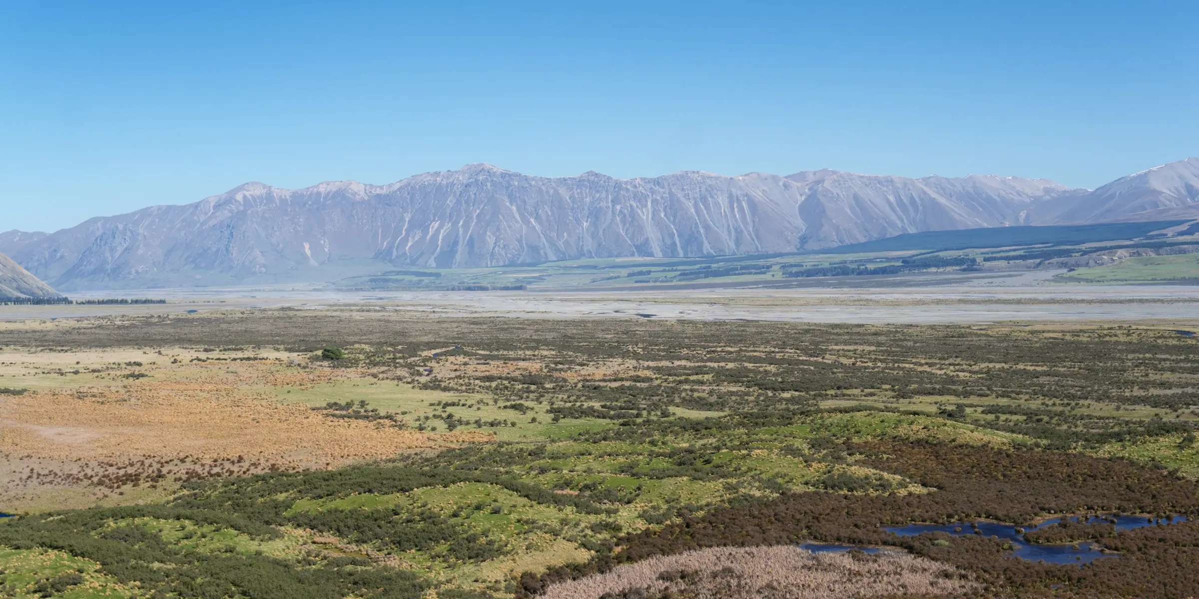 High-country wetlands and tussock flats stretching to a long range of bare mountains under a clear blue sky.