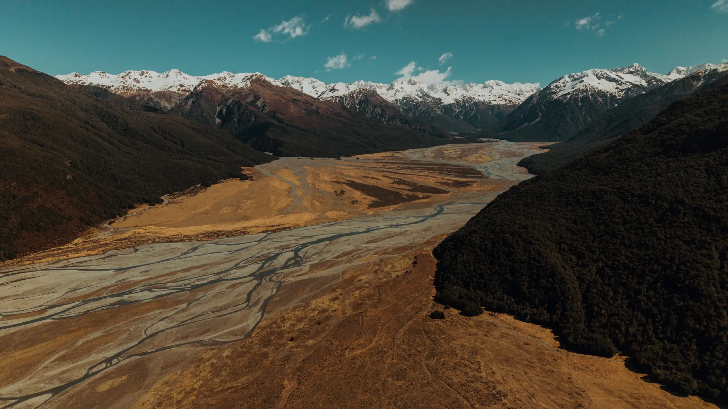 Aerial view of a wide braided riverbed, grey channels across golden shingle flats, running between dark forested slopes towards snow-capped mountains.