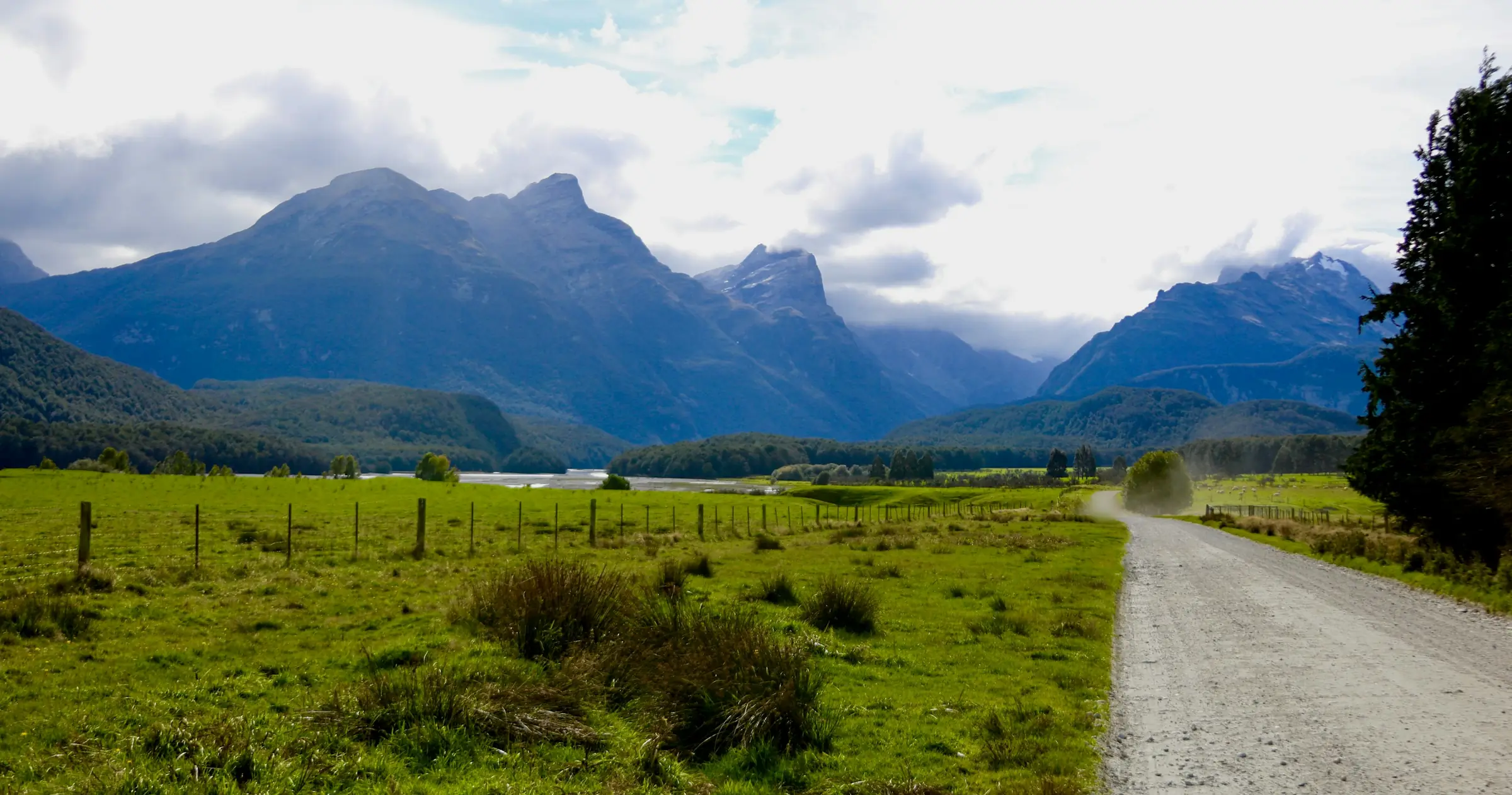 A gravel road running past green paddocks and a wire fence towards forested hills and high mountains under a cloudy sky.